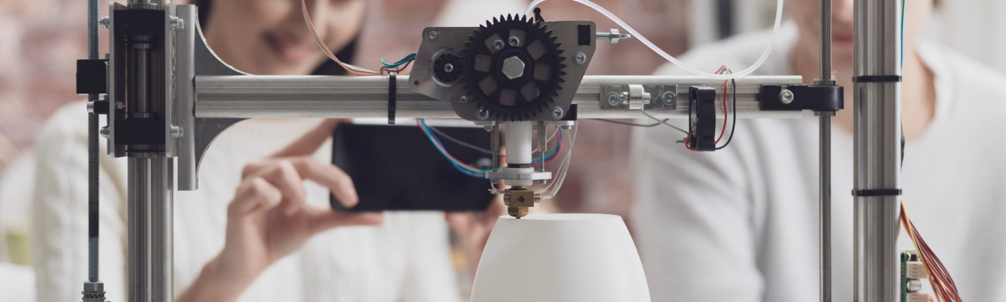 Engineering students printing prototype models using a 3D printer, the girl is taking pictures with a smartphone and sharing online