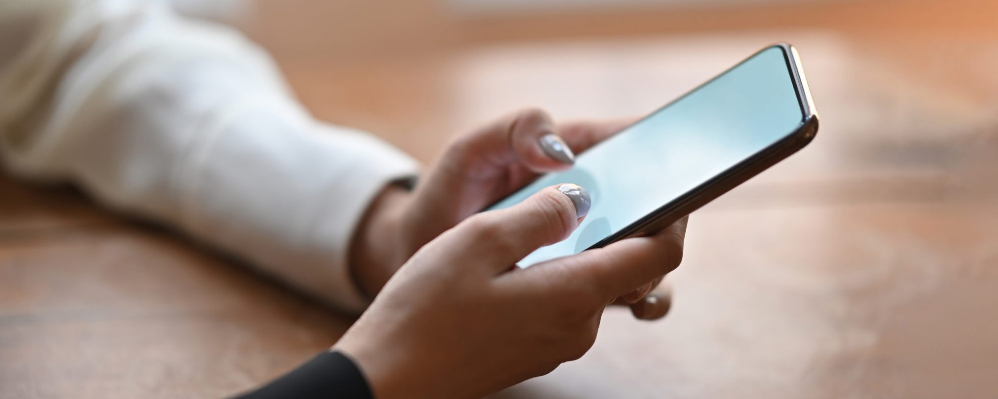 Cropped of woman's hands holding a white blank screen smartphone while sitting and relaxing at the wooden working desk over comfortable living room windows as background.