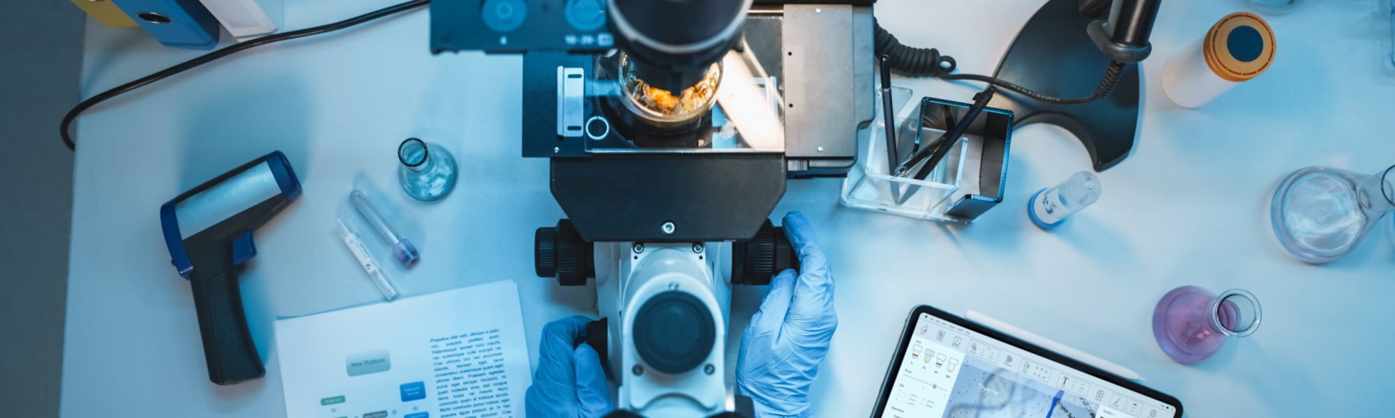 Top Down View of a Medical Research Scientist in Blue Rubber Gloves Working Behind Table in a Modern Laboratory. Doctor is Using Modern Microscop for Sample Analysis and Digital Tablet to Save Data.